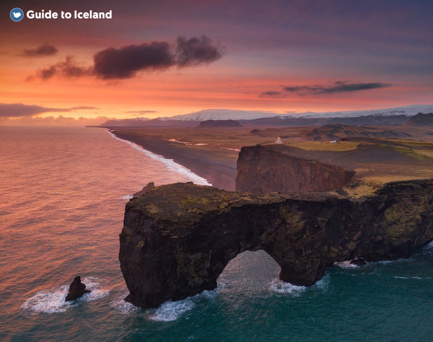 Vista a&eacute;rea de los acantilados y el arco marino de Dyrholaey al atardecer en la Costa Sur de Islandia, con el glaciar Solheimajokull a lo lejos y la luz dorada ba&ntilde;ando las playas de arena negra y el Oc&eacute;ano Atl&aacute;ntico.