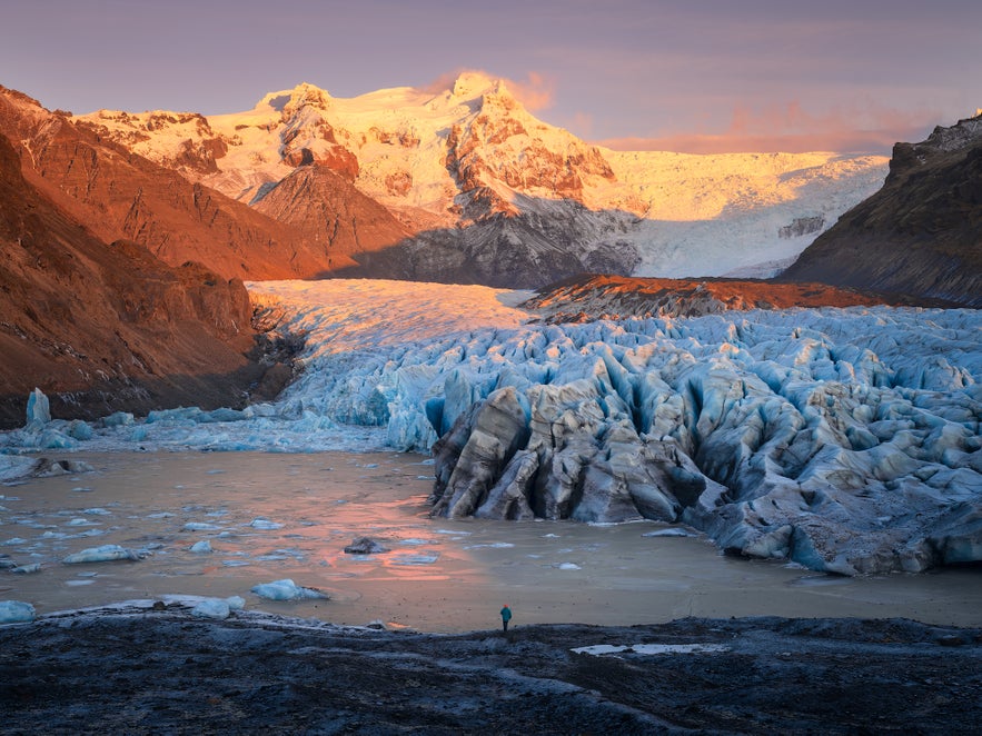 Panoramic view of Svinafellsjokull Glacier in Iceland during sunset, with jagged blue ice formations in the foreground, glowing snow-capped mountains in the distance, and a lone figure standing by the glacial lagoon.