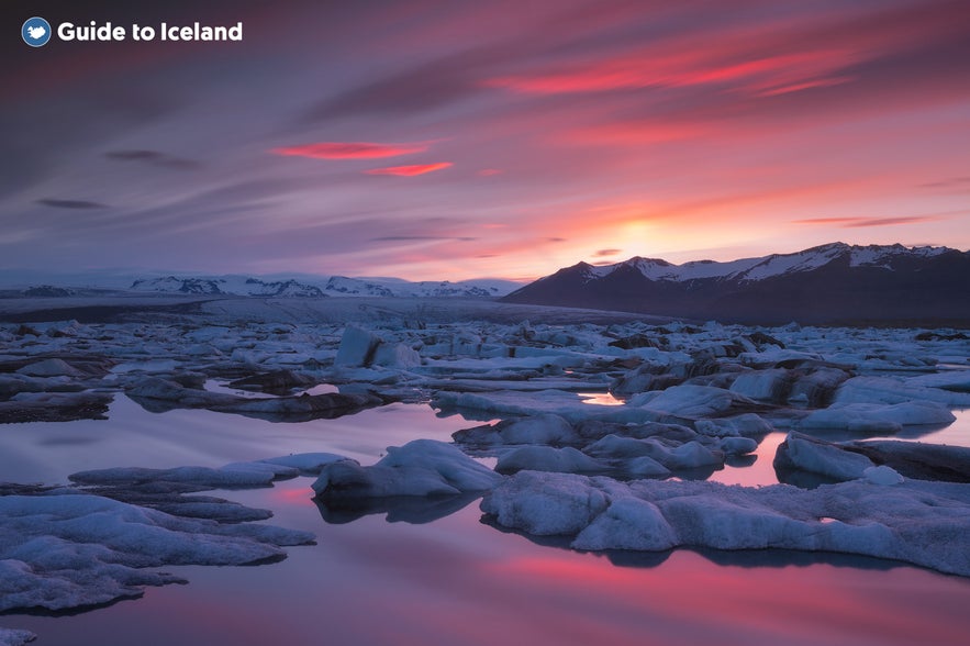 Stunning view of Jokulsarlon Glacier Lagoon at sunset, with icebergs floating on calm reflective water under a dramatic sky glowing in hues of red, orange, and purple.