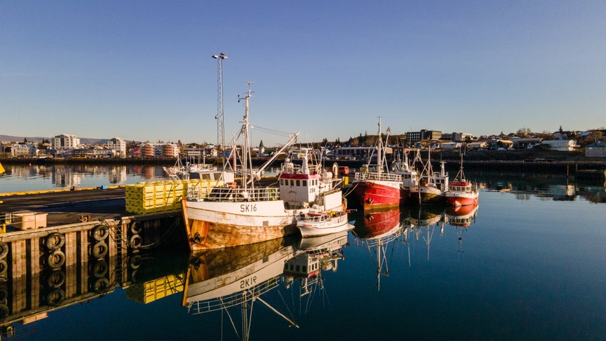 Traditional fishing boats moored at Hafnarfjordur Harbor with a backdrop of colorful buildings and clear blue sky in the Capital Area of Iceland.