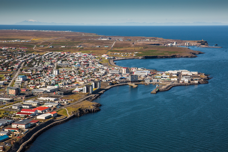  Aerial view of the town center and harbor in Keflavik, with colorful buildings, waterfront piers, and open landscapes stretching toward the horizon.