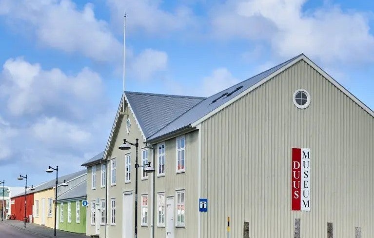 Street view of the Duus Museum in Keflavik, featuring a pale wooden building with red and white signage.