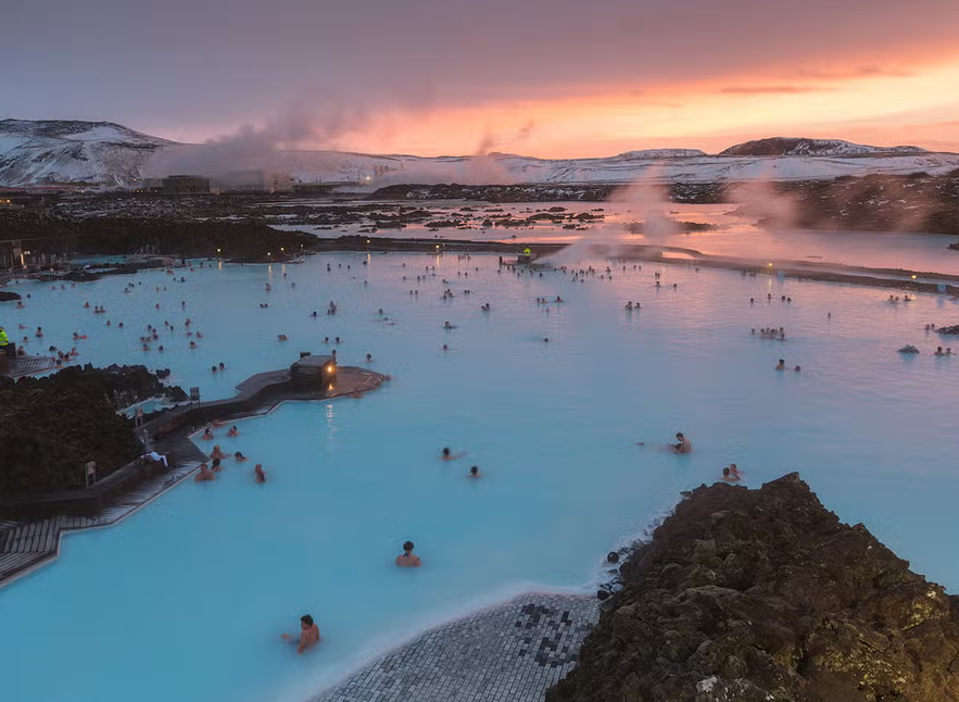 Visitors relaxing in the milky blue waters of the Blue Lagoon at sunset, with steam rising against a snowy volcanic landscape.