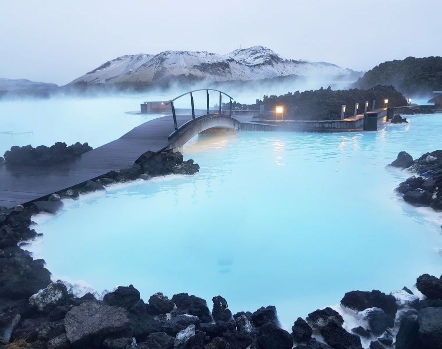 Bridge over the steaming milky blue waters of the Blue Lagoon, surrounded by black lava rocks and snow-dusted hills.