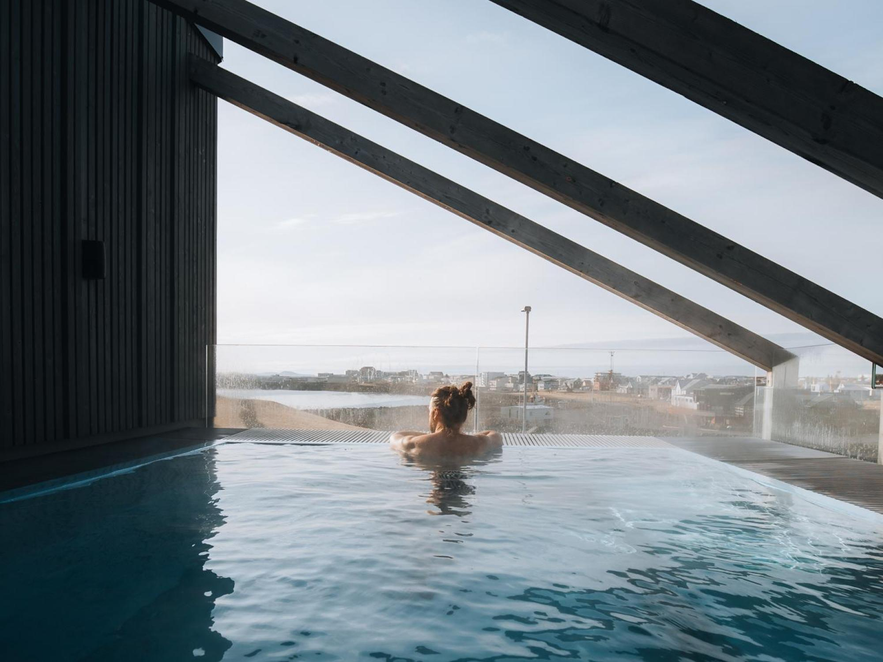 Person relaxing in a rooftop geothermal pool with panoramic views over Keflavik town and coastline.