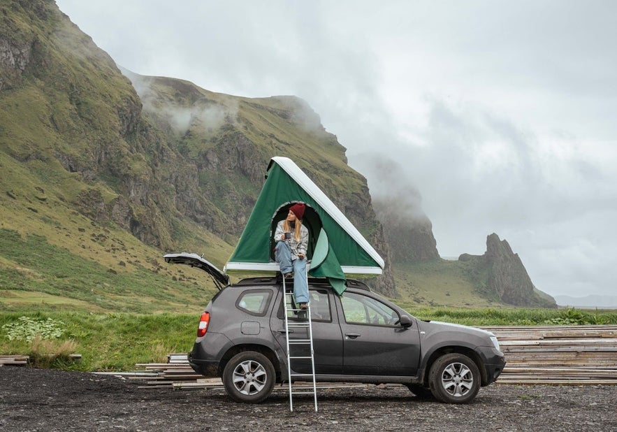 Woman camping in Iceland in a rooftop tent with dramatic mountain views in the background.