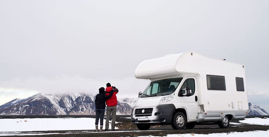 Couple standing beside a campervan in winter, looking at snow-covered mountains.