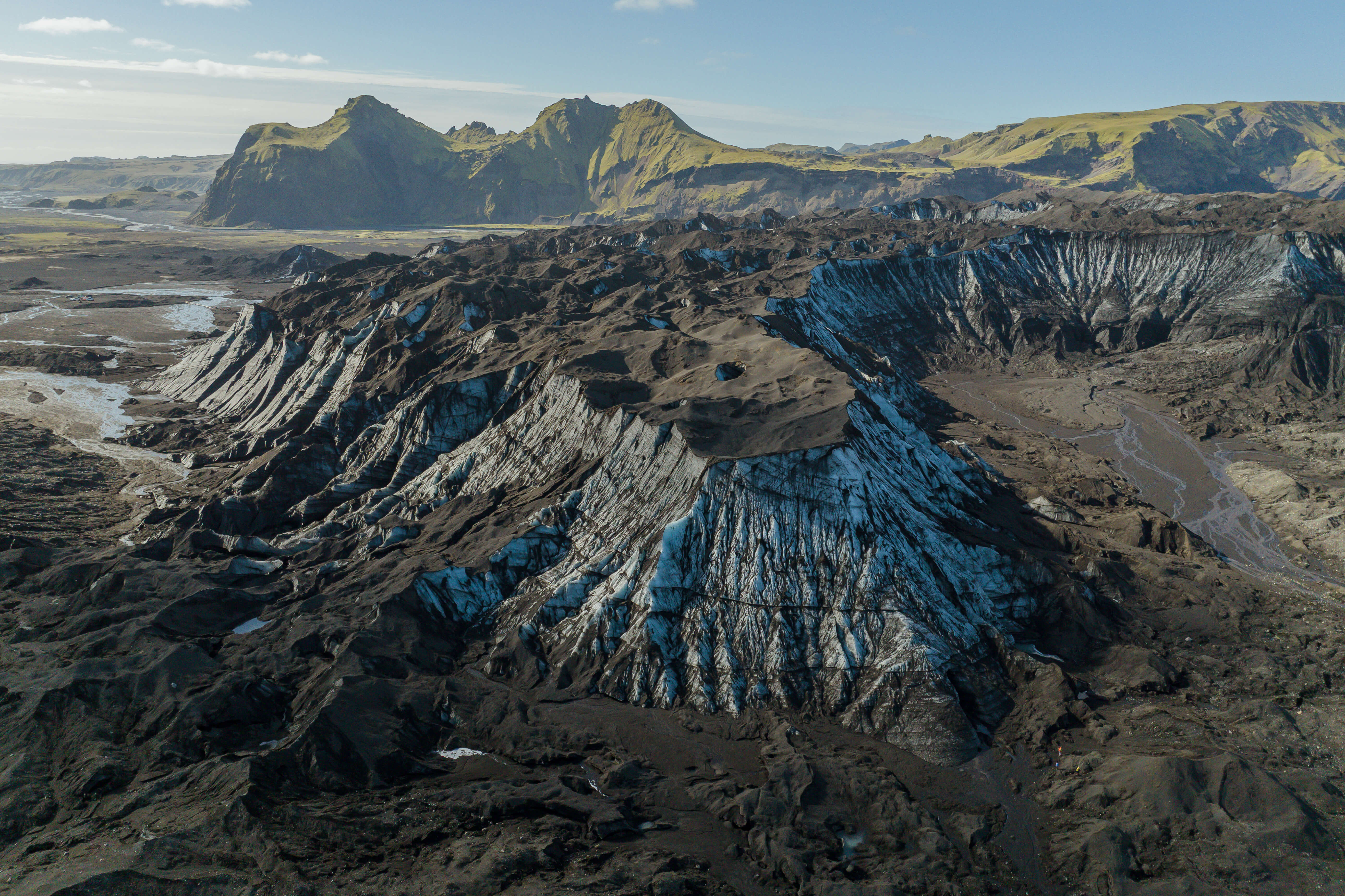 Aerial view of Katla Volcano’s glacier outlet with sharp ice ridges covered in volcanic ash, surrounded by highland cliffs.