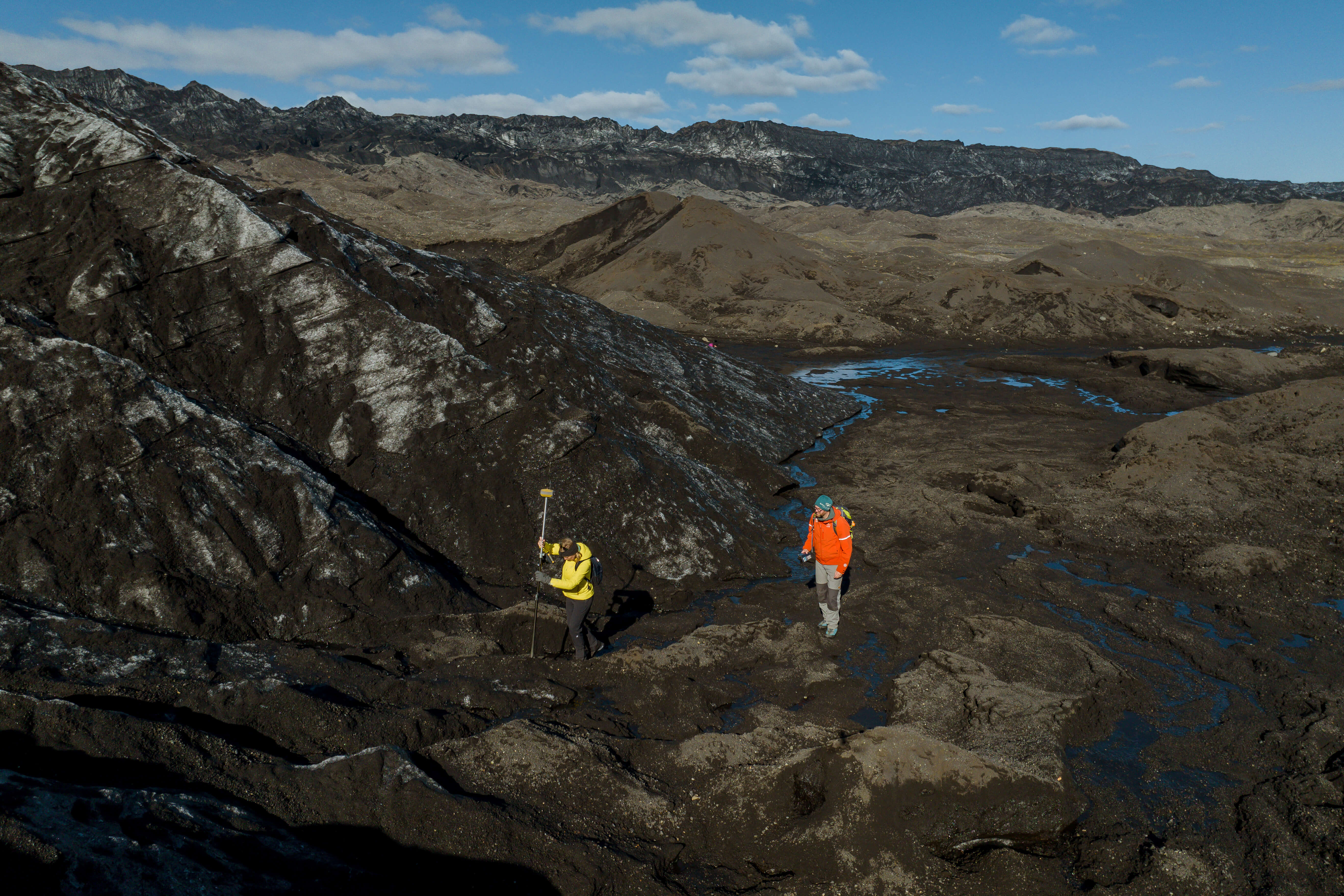 Two researchers in bright gear conduct glacier mapping near Katla using GPS equipment on dark, ice-streaked terrain.