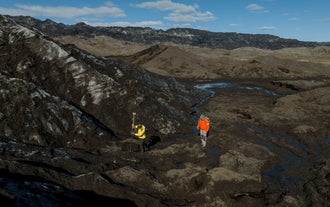 Two researchers in bright gear conduct glacier mapping near Katla using GPS equipment on dark, ice-streaked terrain.