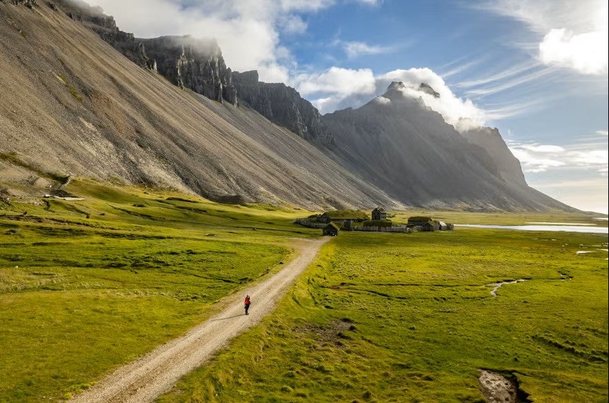 Cyclist riding along a gravel path through the lush green plains beneath Vestrahorn mountain in East Iceland, with dramatic cliffs and Viking village film set in the background.