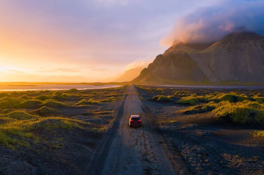 Sonnenuntergang: Ein Auto fährt auf einer schwarzen Sandstraße in Richtung Vestrahorn in Island. Zu sehen sind goldenes Licht und dramatische Wolken entlang der Ringstraße.