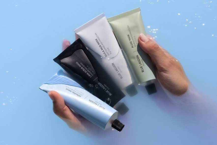 Manos sosteniendo una selecci&oacute;n de mascarillas faciales Blue Lagoon Iceland, incluyendo mineral, lava, algas y s&iacute;lice, sobre el agua geot&eacute;rmica azul lechoso de la Laguna Azul.