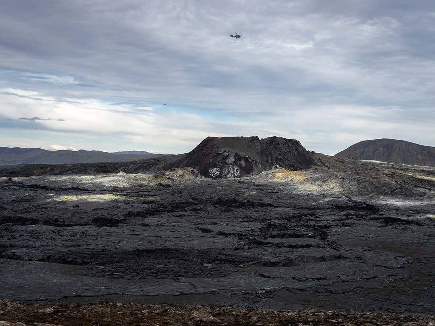 Een helikopter vliegt boven de ruige lavavelden en krater van de vulkaan Fagradalsfjall in IJsland, met het indrukwekkende vulkanische landschap van het schiereiland Reykjanes onder een bewolkte hemel.
