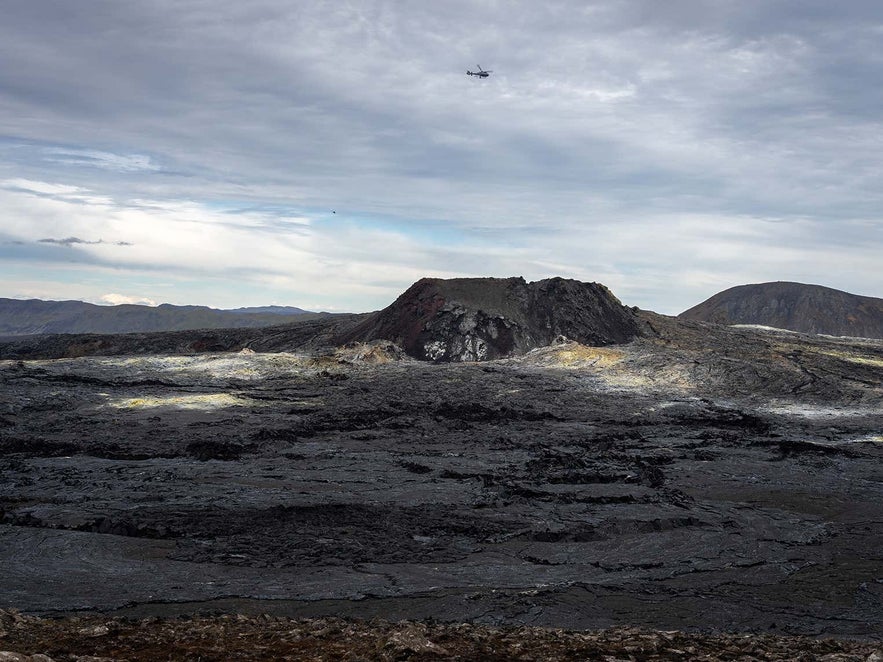 Helicopter flying over the rugged lava fields and crater of Iceland's Fagradalsfjall volcano, showcasing the dramatic volcanic landscape of the Reykjanes Peninsula under cloudy skies.