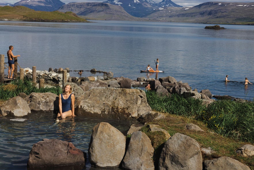 People relaxing in geothermal hot springs at Hvammsvik by the ocean in Iceland, with paddleboarders and swimmers enjoying the fjord under a clear summer sky, surrounded by mountains.