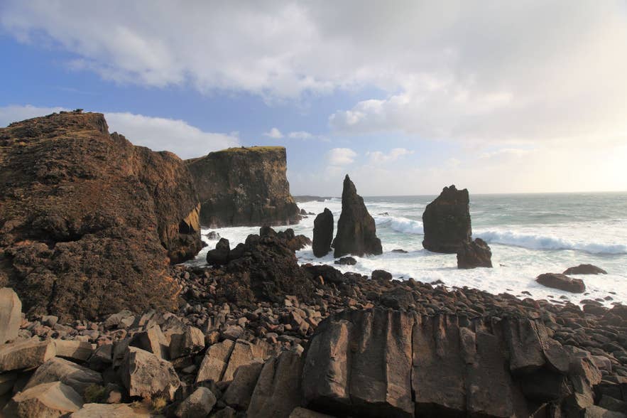 Indrukwekkende zeestapels en ruige zwarte vulkanische kliffen langs de kust van het schiereiland Reykjanes in IJsland, met krachtige golven die onder een deels bewolkte hemel tegen de kust slaan.
