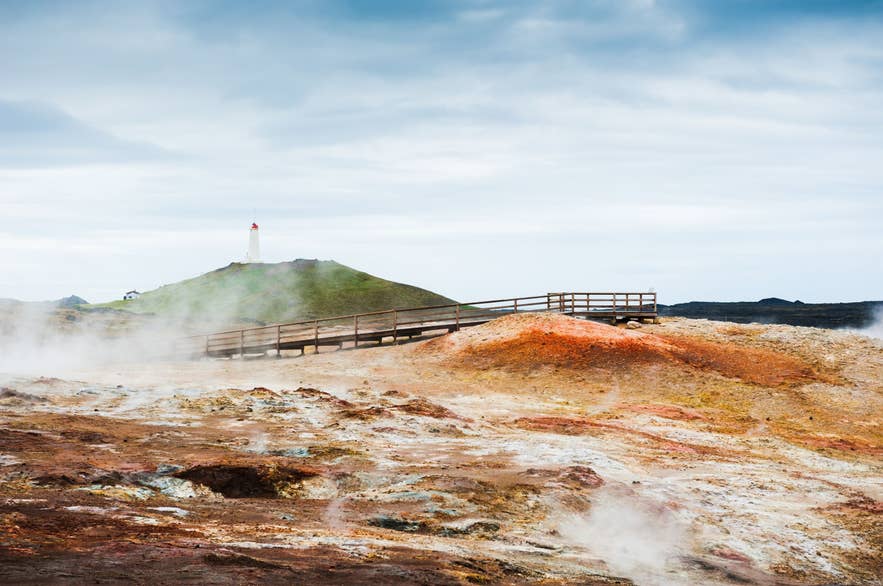 Steaming geothermal landscape with colorful soil at Gunnuhver hot springs in Reykjanes, Iceland, featuring a wooden boardwalk and Reykjanesviti lighthouse in the background under a cloudy sky.