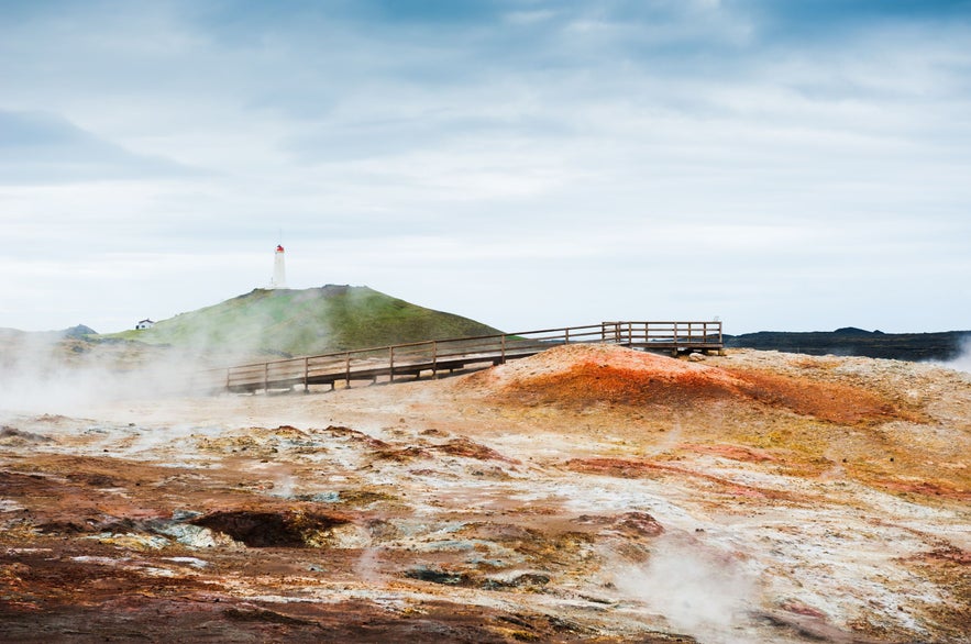 Dampfende geothermale Landschaft mit farbenpr&auml;chtigen Mineralablagerungen an den Gunnuhver-Hei&szlig;quellen auf der Reykjanes-Halbinsel in Island. Ein h&ouml;lzerner Steg f&uuml;hrt durch das Gebiet, w&auml;hrend im Hintergrund der Leuchtturm Reykjanesviti unter einem bew&ouml;lkten Himmel zu sehen ist.