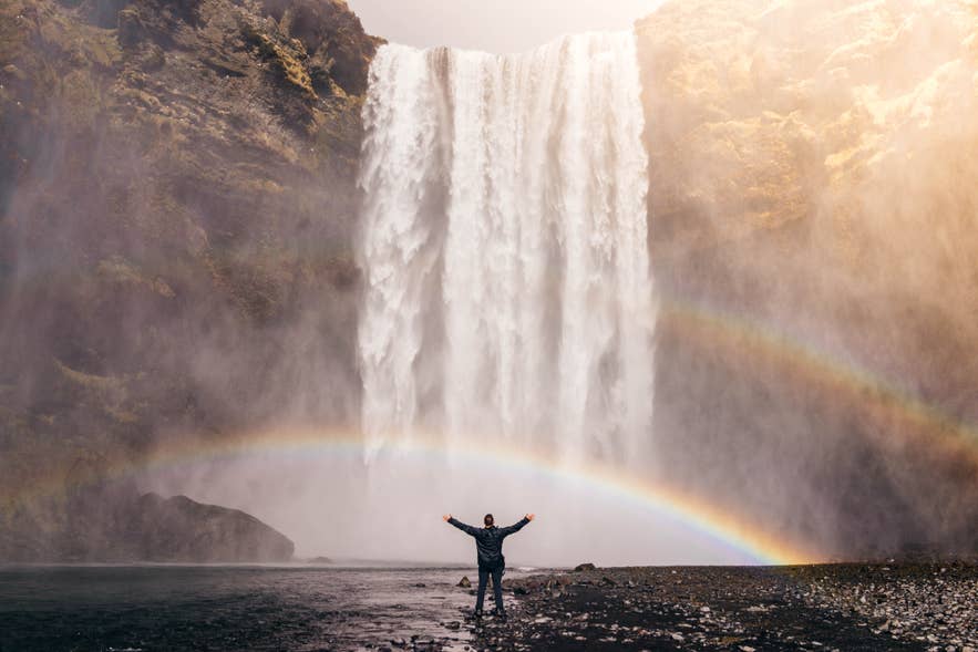 Traveler at Skogafoss waterfall in Iceland with vibrant double rainbow and mist in dramatic sunlight