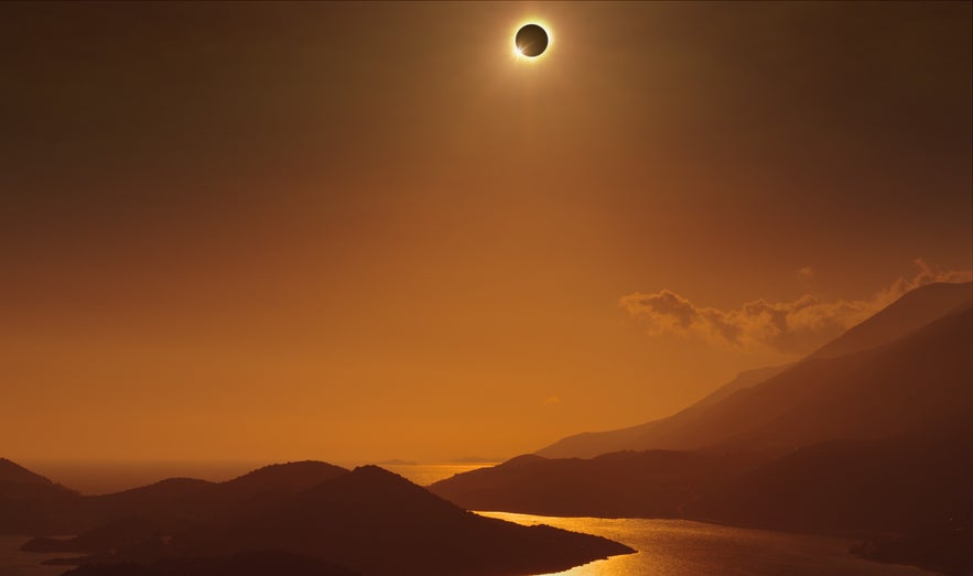A total solar eclipse above a coastal mountain range at sunset casts a warm glow over the sea and hills.
