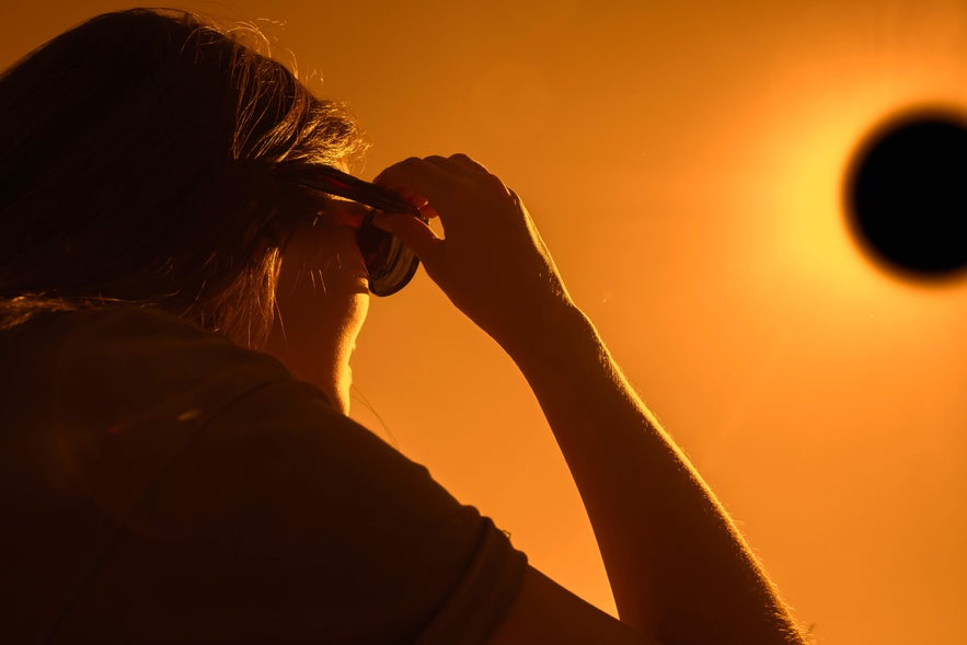 A person is using eclipse glasses to view the Sun during a solar eclipse under a golden-orange sky.