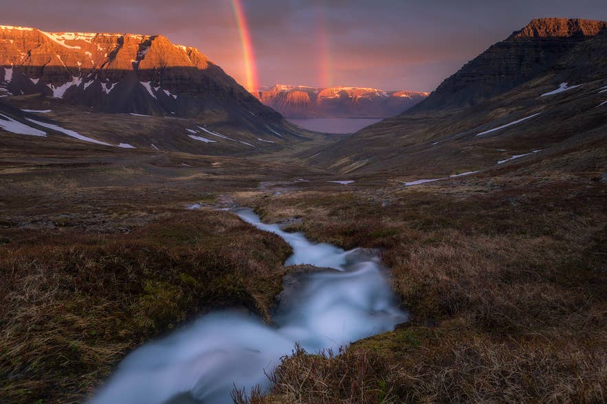 A double rainbow appears over Iceland's Westfjord at sunset, with snow-patched mountains and a flowing stream.