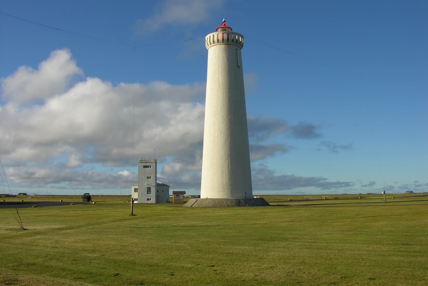 El segundo Faro de Gardskagi, una torre blanca alta con la parte superior roja, se alza sobre un prado bajo un cielo azul con nubes dispersas.