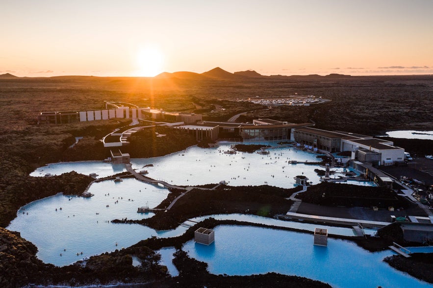 The Blue Lagoon geothermal spa glows under the sunset, with people bathing in milky-blue water surrounded by black lava fields.