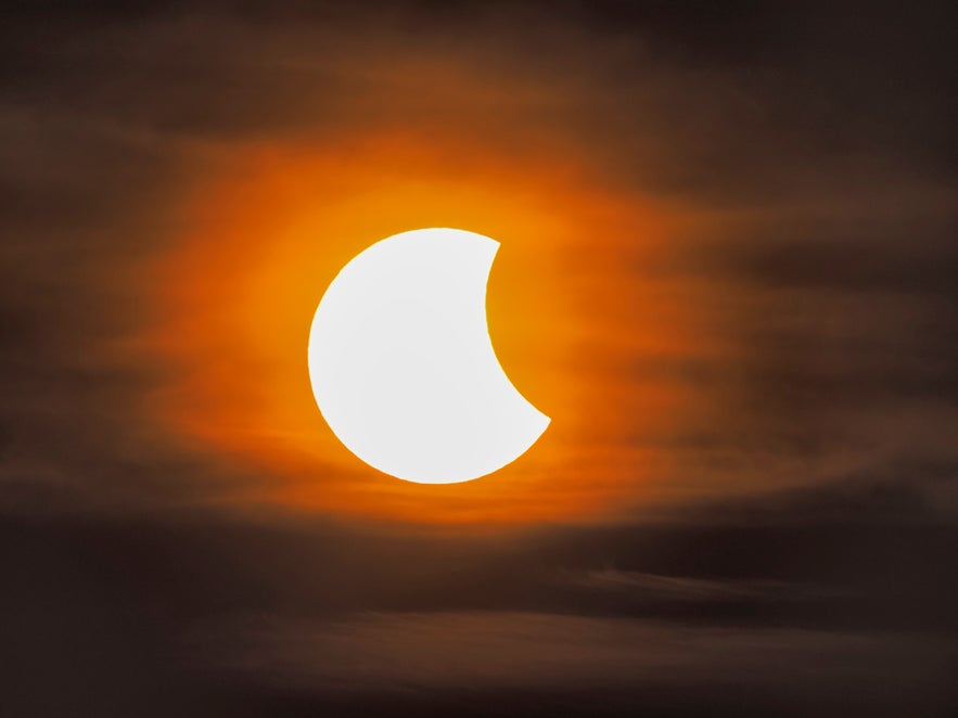 A partial solar eclipse reveals a crescent Sun glowing through thin clouds in an orange sky.