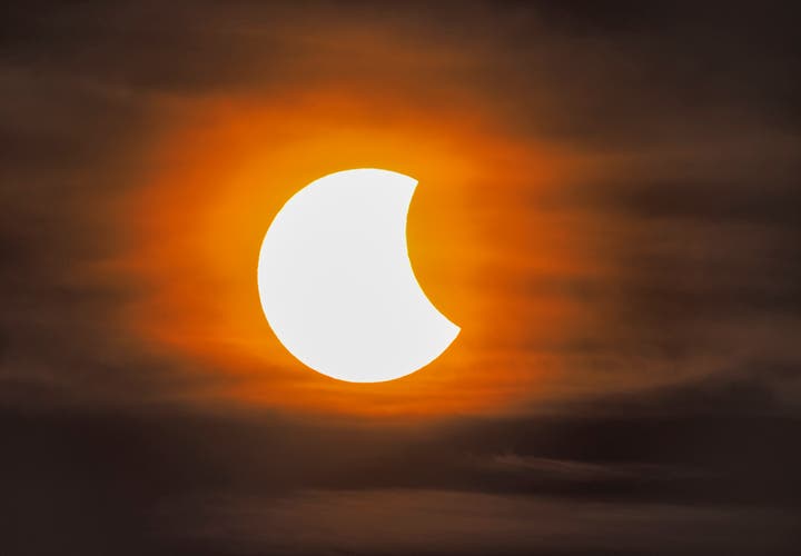 A partial solar eclipse reveals a crescent Sun glowing through thin clouds in an orange sky.