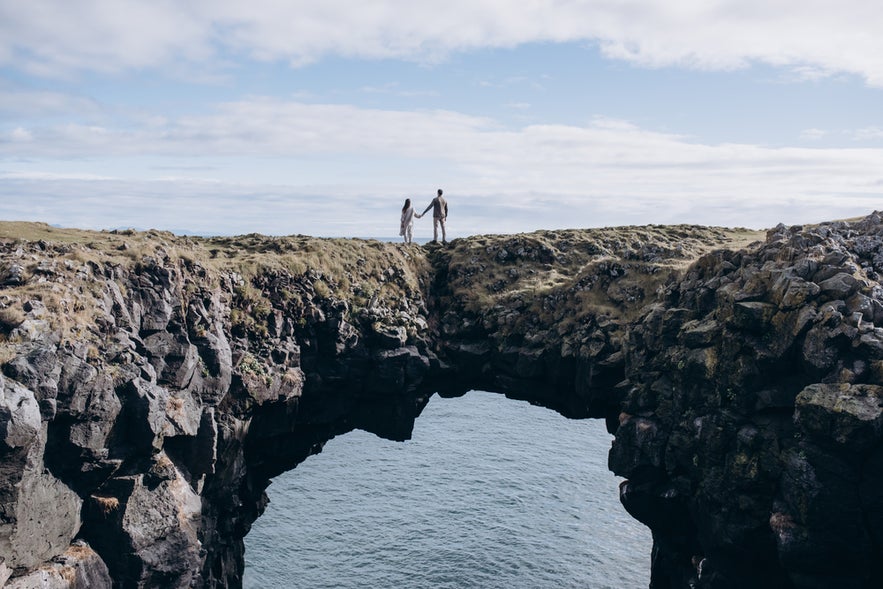 A couple holds hands at a stone gate on Iceland's Snæfellsnes, gazing at the ocean beneath a black volcanic cliff