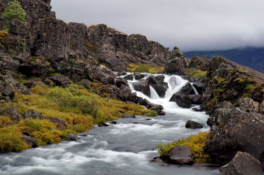 Drekkingarhylur Pool and waterfall in Thingvellir National Park, Iceland, during autumn, with clear cascading water, black volcanic rocks, mossy cliffs, and dramatic cloudy sky on the Golden Circle route.
