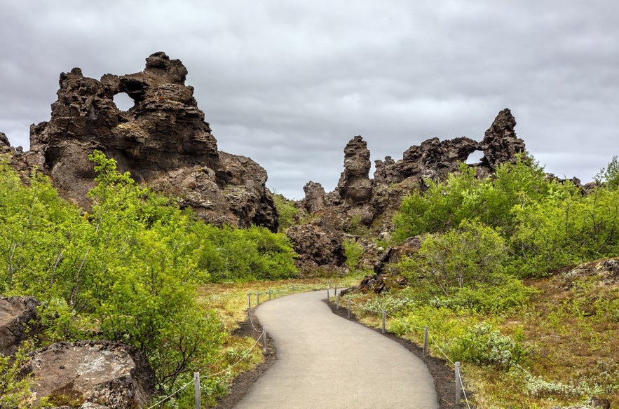 Walking path through Dimmuborgir lava formations near Lake Myvatn in North Iceland, with dramatic volcanic rock pillars, natural arches, green summer vegetation, and cloudy sky.