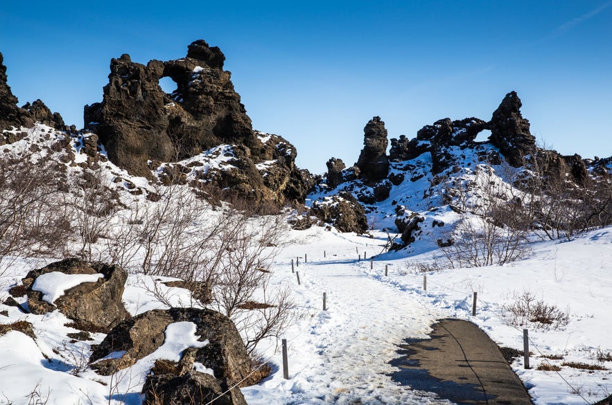 Winter walking path through Dimmuborgir lava formations near Lake Myvatn in North Iceland, with volcanic rock arches, snow-covered landscape, bare trees, and clear blue sky.