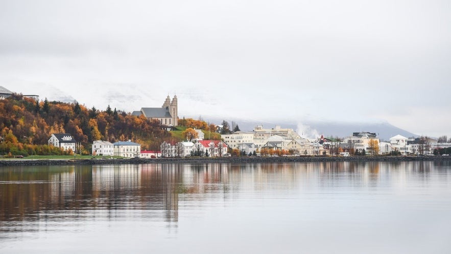 Akureyri town in North Iceland during autumn, with colorful buildings, Akureyrarkirkja church, forested hillside, and calm fjord reflections under soft overcast sky.