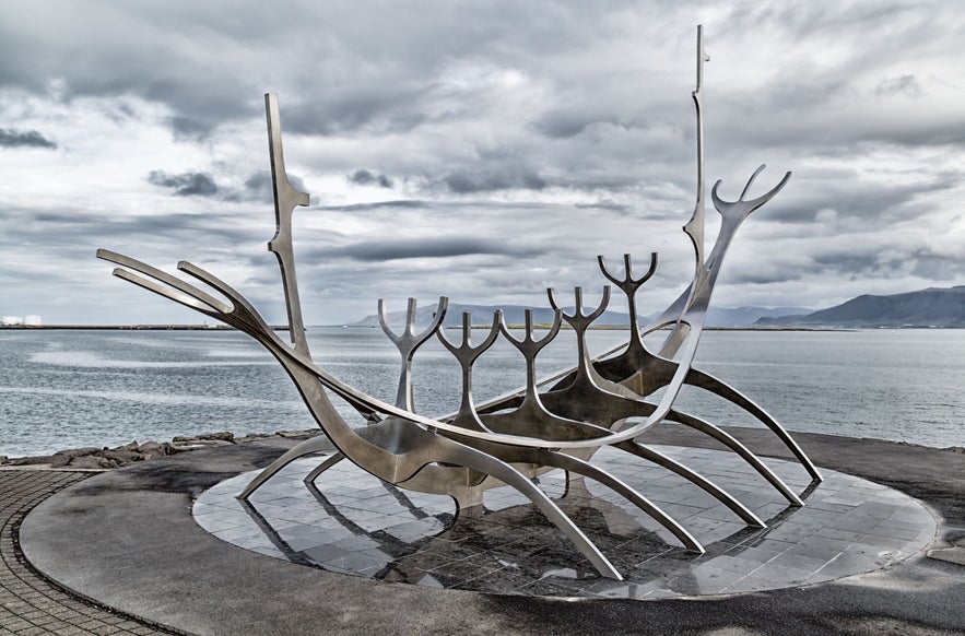 Sun Voyager sculpture in Reykjavik, Iceland, with modern stainless steel design, overlooking Faxaflói Bay, distant mountains, and dramatic cloudy sky.