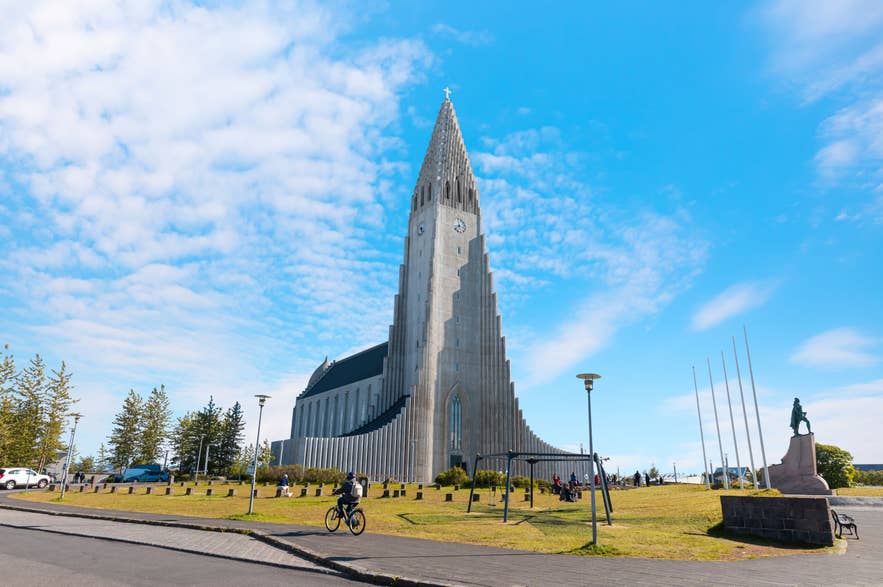 아이슬란드 레이캬비크에 위치한 할그림스키르캬(Hallgrimskirkja) 교회. 높은 콘크리트 첨탑, 독특한 모더니즘 건축 양식, 레이프 에릭손 동상, 푸른 여름 하늘, 그리고 교회 광장에 모인 방문객들의 모습.