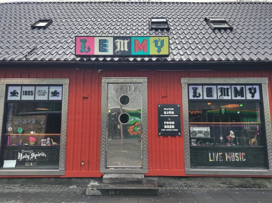 Exterior of Lemmy Bar in Reykjavik, Iceland, with red wooden facade, colorful signage, large windows, neon interior lights, and live music advertisement.