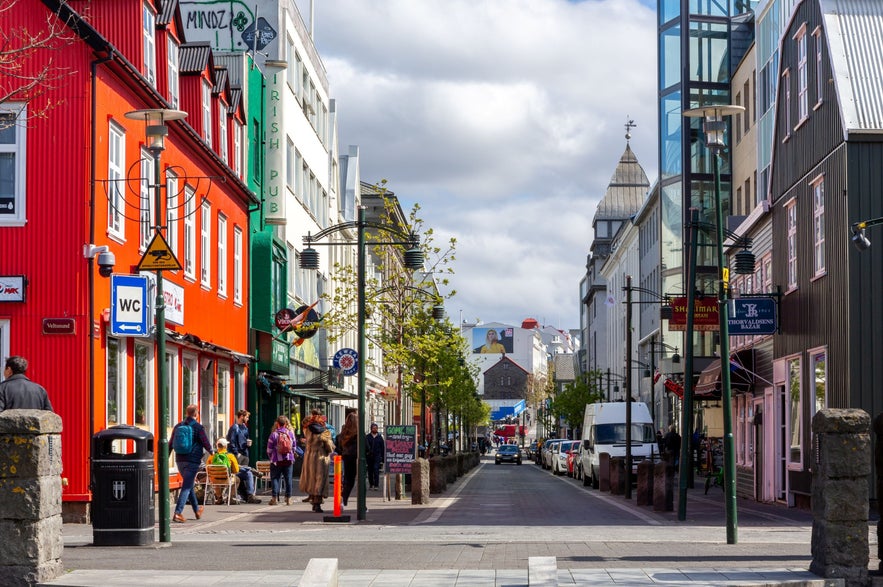 Austurstraeti street in downtown Reykjavik, Iceland, with shops, restaurants, outdoor cafes, pedestrians, cars, and Laugavegur Street in the distance under summer sky.