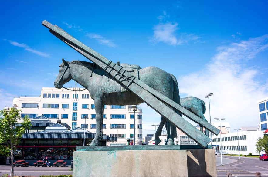 Horse statue at Hlemmur Square in Reykjavík, Iceland, with modern city buildings, bright blue summer sky, and the nearby Hlemmur Mathöll food hall in background.