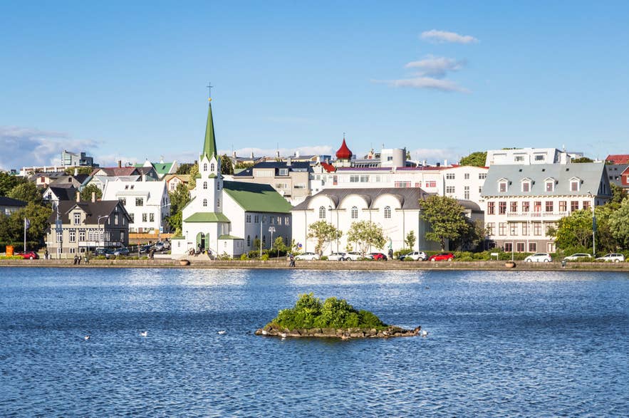 Das Stadtbild von Reykjavík, gesehen vom See Tjörnin in Island, mit der Frikirkjan-Kirche, den historischen Gebäuden der Innenstadt, dem ruhigen blauen Wasser, einer kleinen Insel und dem klaren Sommerhimmel.