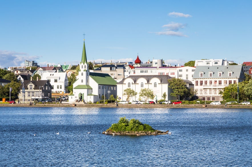 Reykjavik cityscape viewed from Tjornin lake, Iceland, with Frikirkjan church, historic downtown buildings, calm blue water, small island, and clear summer sky.
