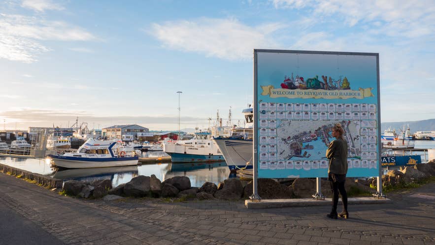 Der alte Hafen von Reykjavik in Island mit Fischerbooten, Yachthafen, einer Hafenkarte, einer Person, die die Karte liest, ruhigem Wasser und sanftem Sommerabendlicht.