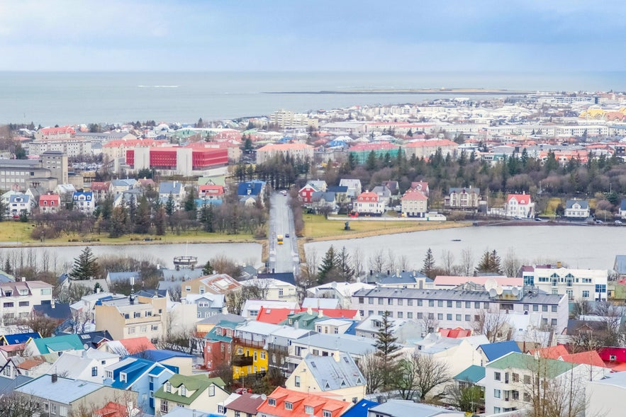 Vesturbaer neighborhood in Reykjavik, Iceland, viewed from above, with colorful rooftops, Tjörnin lake, residential houses, trees, and distant ocean horizon.