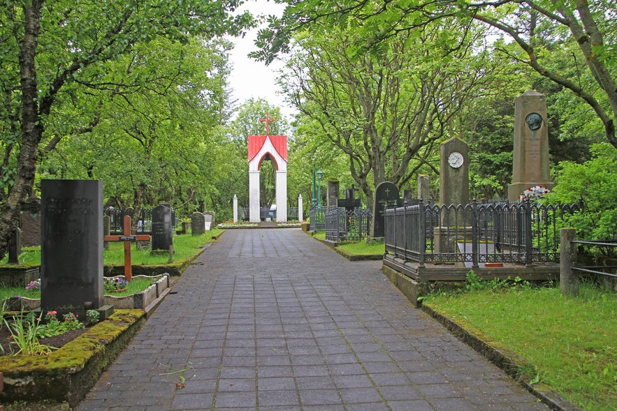 Holavallagardur cemetery in Reykjavik, Iceland, with gravestones, mossy tombs, trees, paved walkway, red-roofed chapel, and peaceful green summer landscape.