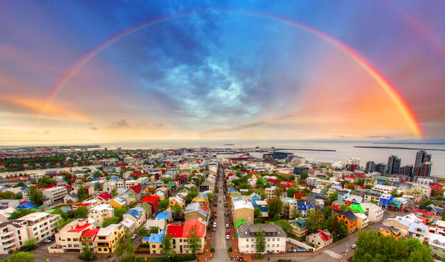 Vibrant aerial view of Reykjavik city center with colorful rooftops and a full rainbow arching over Faxafloi Bay, Iceland.