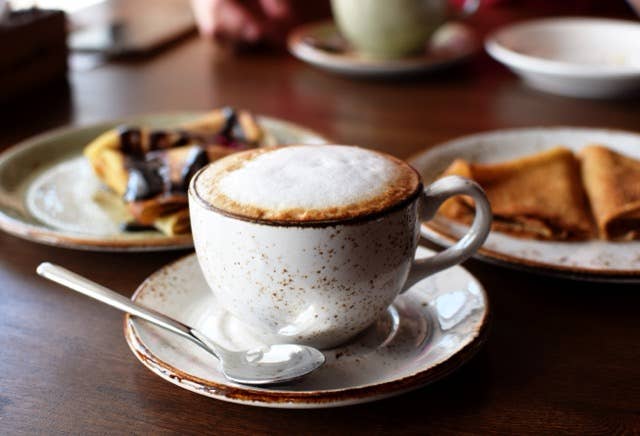A cup of frothy cappuccino with two plates of bread and pastry in the background