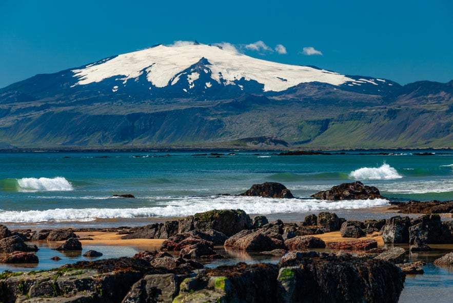 Snaefellsjokull glacier and volcano on Snaefellsnes Peninsula in Iceland with black lava rocks, golden beach, and turquoise ocean waves in summer.