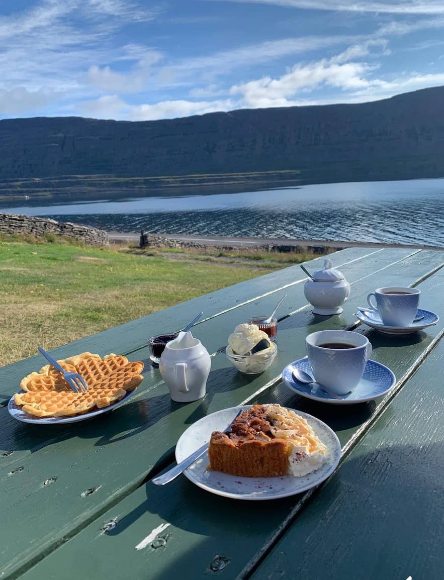 Plates of waffles and pies and cups of coffee on a green table by the fjord at Litlibaer, a coffee shop in the Westfjords